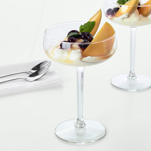 Dos elegantes vasos de tallo rellenos de fruta y crema de postre, mostrados en una mesa blanca con cucharas cerca.