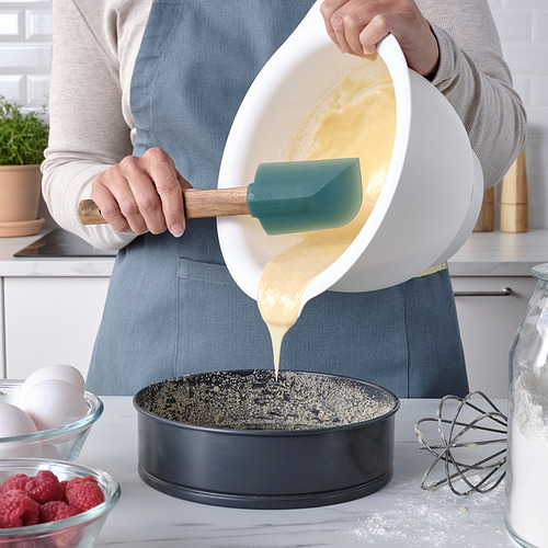 Una persona vierte la masa de un tazón blanco con una espátula verde en una sartén redonda, rodeada de ingredientes para hornear en una encimera de la cocina.