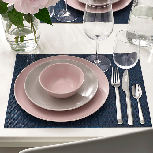 Mesa con platos, cuencos y cubiertos rosas sobre manteles individuales azules. Incluye copas de vino y un jarrón con flores rosas.