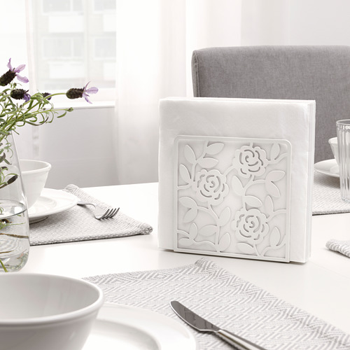 Un servilletero blanco, LIKSIDIG, con un diseño de rosas, mantiene las servilletas en posición vertical sobre una mesa con un cuenco blanco, cubiertos y un jarrón con flores.