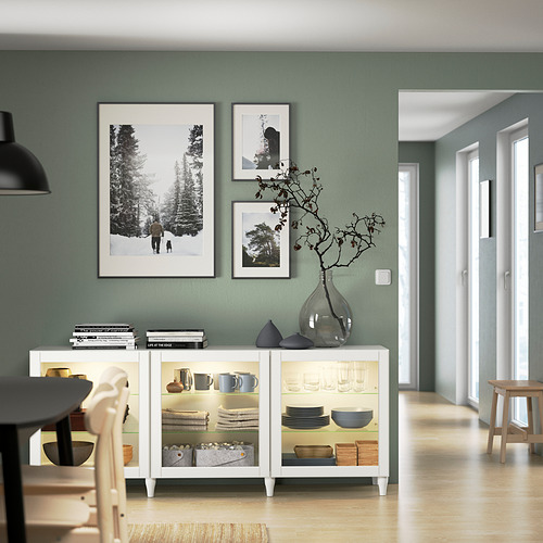 Aparador blanco con puerta de cristal BESTÅ con platos, decoración y jarrón. Muro verde.