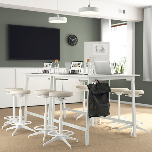 Configuración de oficina moderna: mesas altas MITTZON con mesas blancas y taburetes beige, pantallas planas, computadoras portátiles y mochilas.