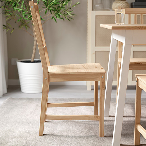 Silla de madera de color marrón claro con respaldo de listones verticales y diseño sencillo, colocada cerca de una mesa blanca con una planta al fondo.