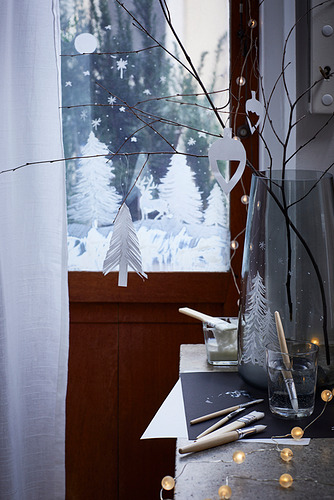 Ventana nevada con jarrón, ramas desnudas y adornos de papel blanco, suavemente iluminados.