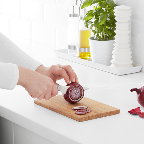 La persona corta la cebolla en rodajas en una tabla de cortar de bambú sobre una encimera blanca, con accesorios de cocina detrás.