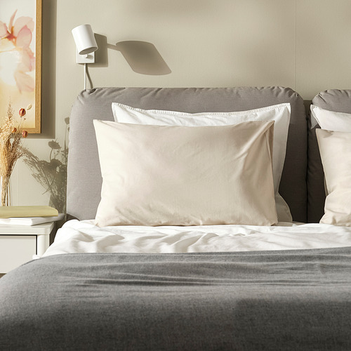 Funda de almohada crema DVALA, algodón suave, forma cuadrada, sobre una cama grande, cabecero gris, diseño sencillo.
