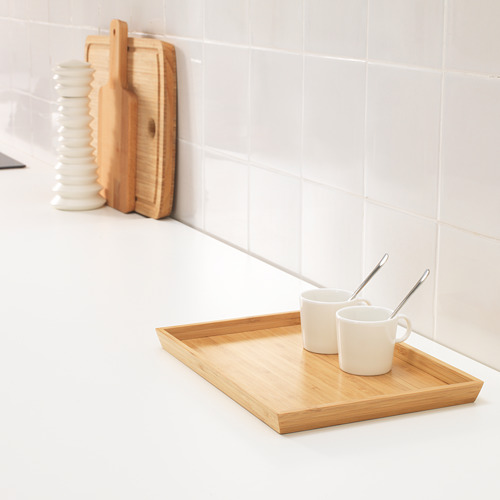 Bandeja de madera con dos tazas y cucharas blancas, colocada sobre una encimera blanca junto a tablas de cortar de madera y una pila de platos blancos.