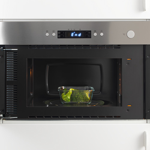 Microondas de acero inoxidable con recipiente de vidrio de brócoli en su interior, mostrando su función de calentar o descongelar alimentos.