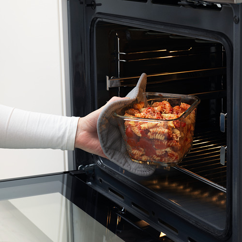 Coloca a mano un plato de vidrio con pasta en un horno negro, mostrando su uso seguro para el horno y su comodidad.