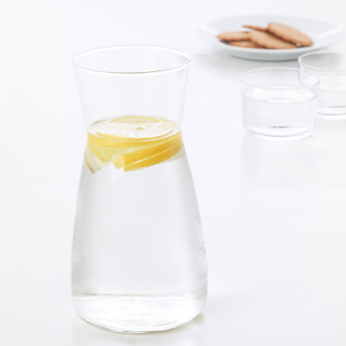 Jarra de vidrio transparente con rodaja de limón en el interior, colocada sobre una superficie blanca cerca de un vaso vacío y un plato con galletas.
