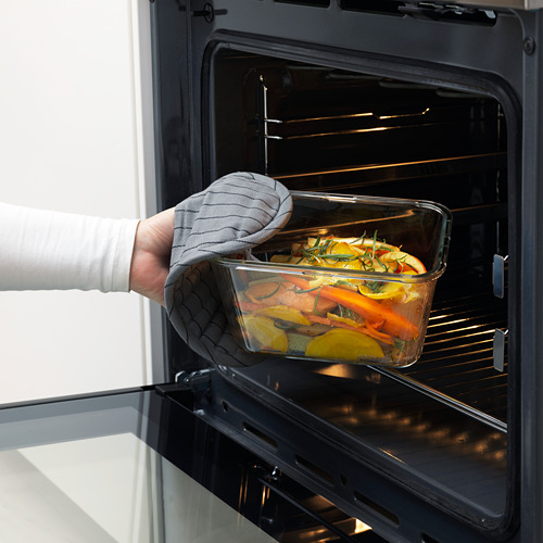 Persona que inserta un recipiente de vidrio para alimentos, lleno de verduras, en un horno abierto, usando un guante de cocina.