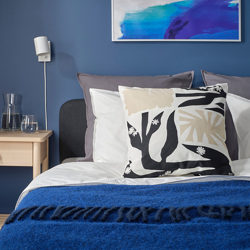 Dormitorio azul con cama floral, almohadas grises, manta azul, mesita de noche de madera, jarra de cristal, arte.