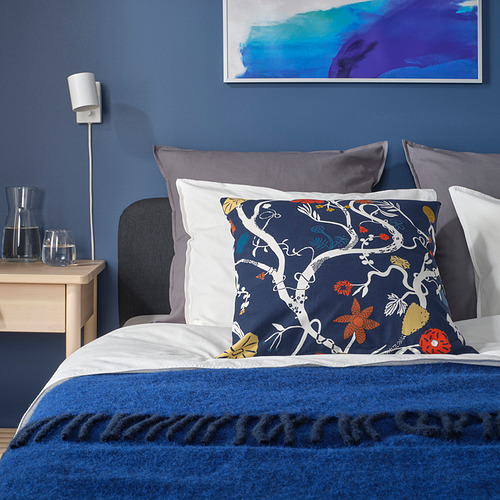 Cojín floral REVFINGERÖRT azul marino sobre elegante cama con almohadas mixtas, manta y mesita de noche de madera.