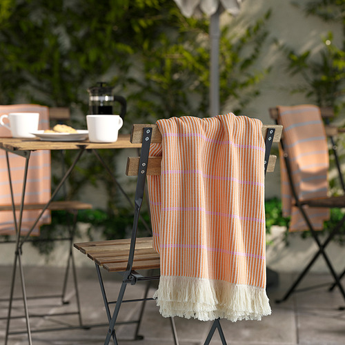 Las mantas de rayas naranjas cuelgan de las sillas de exterior, rodeadas de mesas plegables con tazas y plantas, dando un ambiente acogedor.