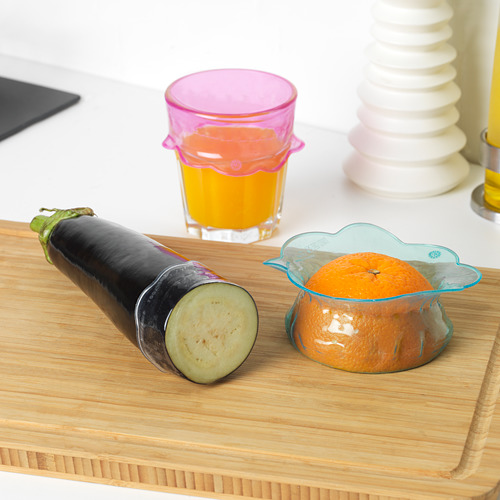 Tabla de cortar de madera con berenjena a medio cortar y una naranja en una funda de silicona junto a un vaso de zumo y jarrones apilables.