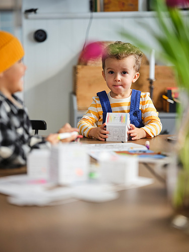 Niños haciendo manualidades en la mesa con materiales, niño con camisa a rayas sostiene modelo, niño con sombrero naranja visible, habitación luminosa con plantas.