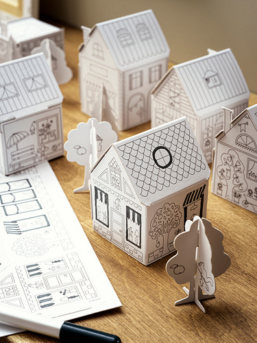 Casas y árboles de cartón plegable en blanco y negro se sientan sobre una mesa de madera, diseñada para que los niños pinten y creen un pequeño pueblo.