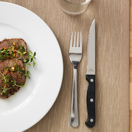 Un cuchillo para carne con hoja dentada y mango negro se coloca junto a un tenedor y un plato con carne cocida adornada con verduras.