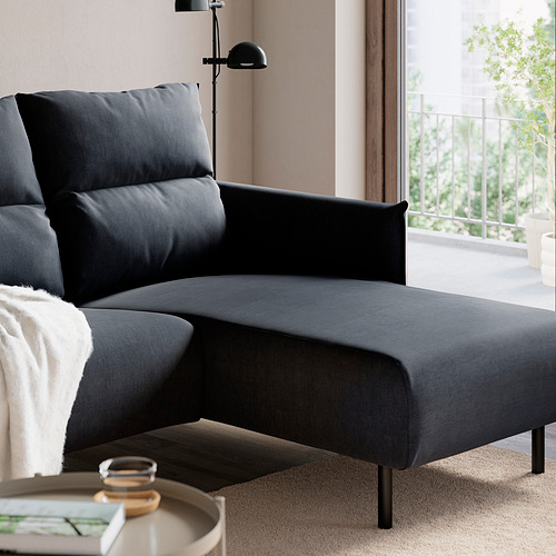Sofá gris con chaise, cojines, patas negras, mesilla con revista y gafas.