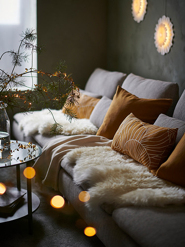 Acogedora sala de estar con un sofá SÖDERHAMN gris, cojines marrones, manta de piel suave y luces festivas.