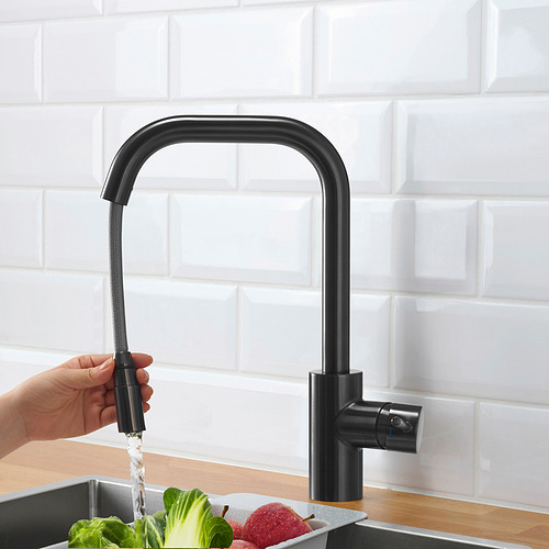 Mano sosteniendo un grifo negro rociando agua en el fregadero con frutas y verduras.