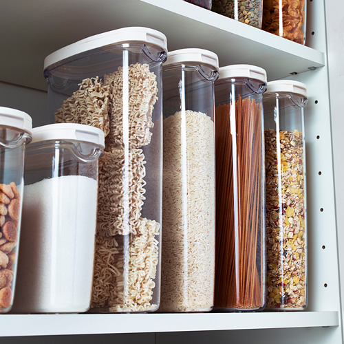 Los recipientes de almacenamiento transparentes con tapas blancas contienen alimentos secos como fideos, arroz y avena. Organizado en un estante blanco para facilitar el acceso.
