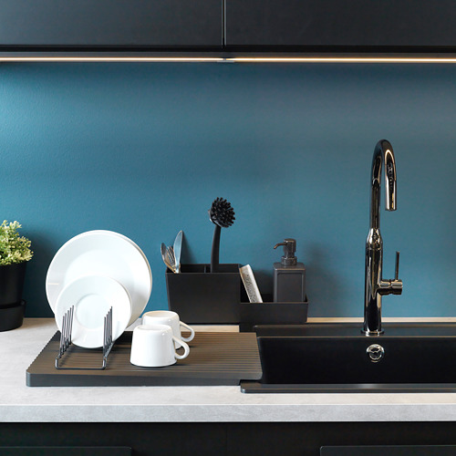 Escena de cocina limpia: platos secándose, tazas en soporte, cepillo, utensilios en soporte, dispensador de jabón en el fregadero con grifo brillante, pared azul.