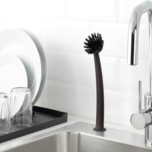 Cepillo de cocina negro con base de ventosa para fregar, apoyado en el fregadero de acero inoxidable cerca del grifo de plata.