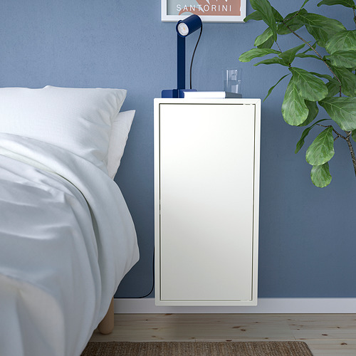 Pequeño armario EKET blanco junto a la cama, pared azul. Sostiene lámpara, planta y libros. Diseño sencillo y moderno.