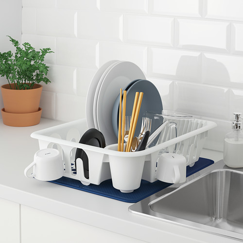 Escurridor de platos blanco, FLUNDRA, sostiene platos, tazones, tazas, cubiertos. Colocado sobre una estera azul cerca del fregadero, con una planta cerca, secando los platos de manera eficiente.
