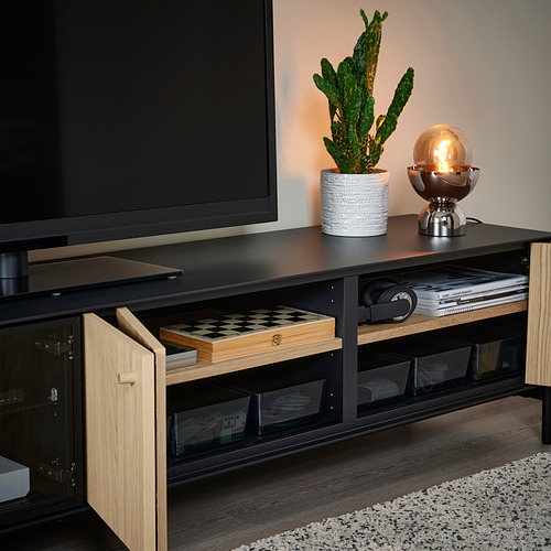 Mueble de TV negro con puertas plegables y estantes de vidrio, que sostiene un televisor, un cactus, una lámpara, auriculares y libros.