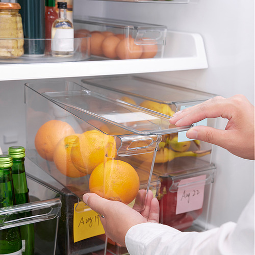 La persona organiza las naranjas en una caja de almacenamiento transparente del refrigerador con tapa. La caja tiene asa incorporada y nota de fecha. Nevera bien organizada con varios artículos.