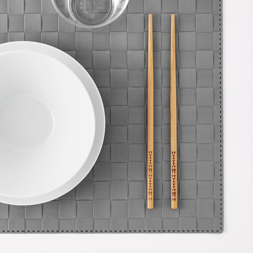 Un par de palillos decorativos de bambú descansan sobre un mantel individual gris, junto a un cuenco blanco y un vaso.