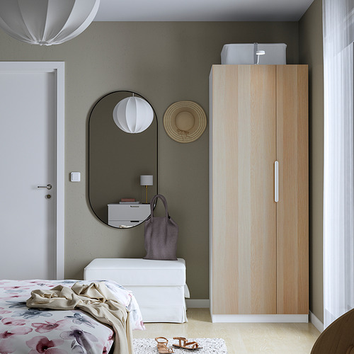 Dormitorio minimalista con armario de madera PAX, tocador blanco y espejo grande. La colcha floral y la lámpara añaden color. El armario ofrece un almacenamiento eficiente.