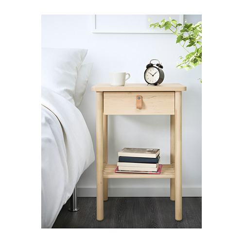 Mesita de noche de madera con cajón y estante, con libros, una taza y un despertador junto a una cama blanca.
