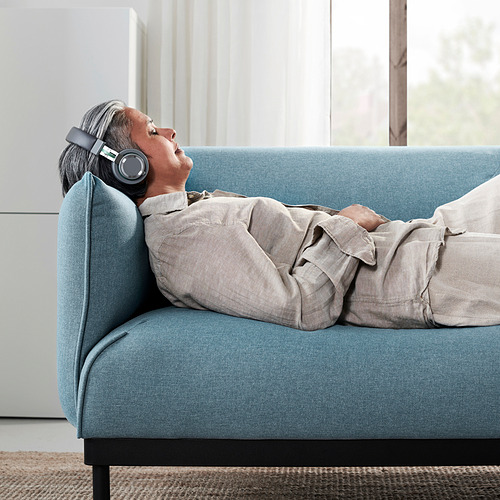 La mujer se relaja en un sofá azul con auriculares, cubierto por una manta beige, en medio de la luz natural.