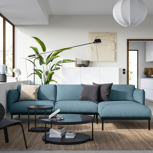 Sala de estar moderna con sofá ÄPPLARYD azul, suelo de madera, mesa de centro de cristal, plantas tropicales y arte abstracto en la pared.