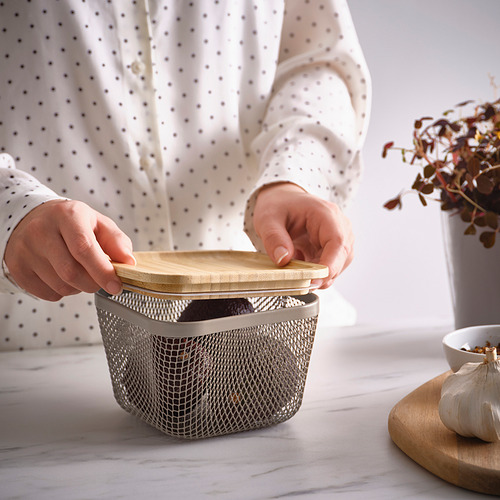 Persona colocando una tapa de bambú en la cesta de almacenamiento de alimentos de malla IKEA 365+ en una encimera de cocina. En las inmediaciones hay un bulbo de ajo y una tabla de cortar.