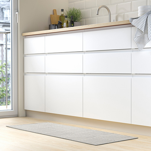 Cocina moderna con armarios blancos. Alfombra de rayas grises y blancas en el suelo, accesorio debajo del fregadero. Encimera de madera con planta verde, botellas y toallas.