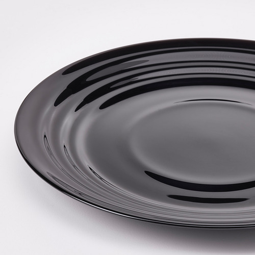 Placa redonda de color negro con un diseño brillante y ondulado sobre una superficie blanca, que refleja la luz, muestra una artesanía de alta calidad y un acabado elegante.