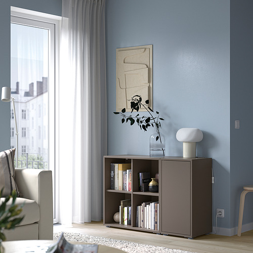Sala de estar moderna con pared azul, arte abstracto, estantería, silla y ventana con cortinas transparentes. Detalle de la unidad de almacenamiento EKET con libros y jarrón.