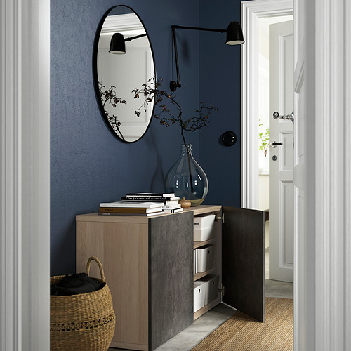 Entrada contemporánea con almacenamiento BESTÅ. La puerta abierta muestra estantes y cajas, pared azul, espejo, jarrón con ramas.