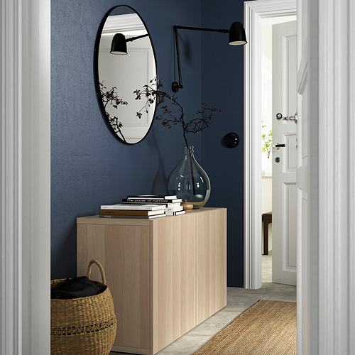 Habitación moderna con pared azul, aparador IKEA BESTÅ de roble, jarrón, libros, cesta, espejo ovalado, lámpara minimalista, almacenaje, puerta a otra habitación.