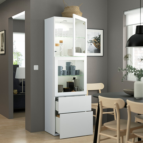 Comedor moderno, un armario blanco BESTÅ con puertas y cajones de vidrio, que exhibe tazas y frascos azules. Junto a una mesa de madera con sillas.