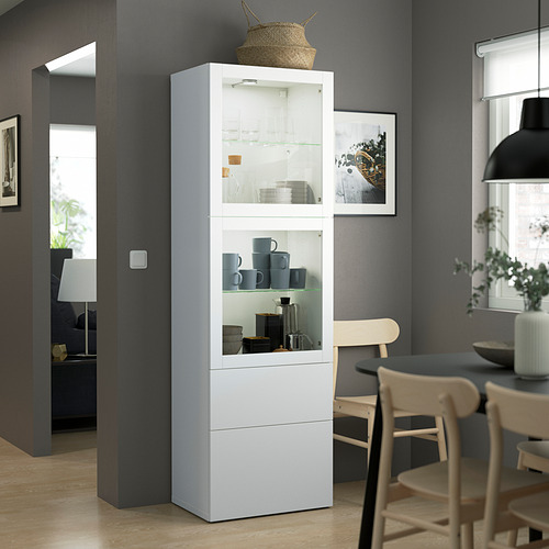 Moderno armario BESTÅ con puertas de cristal, tazas azules, cajones, en una habitación bien iluminada.
