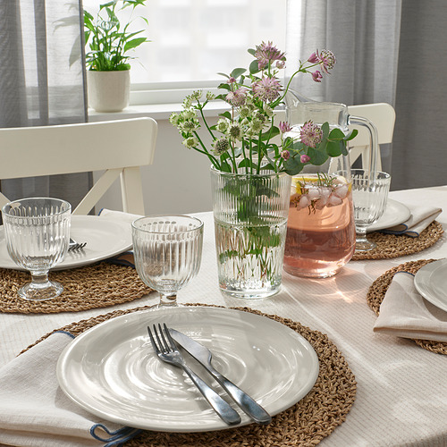 Mesa con platos blancos, cubiertos, manteles individuales tejidos, vasos y jarrones con flores dispuestos sobre un mantel beige, listos para comer.