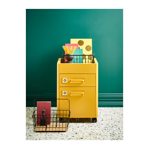 El archivador IDÅSEN amarillo brillante con ruedas contiene coloridos suministros de oficina, de pie contra una pared verde.