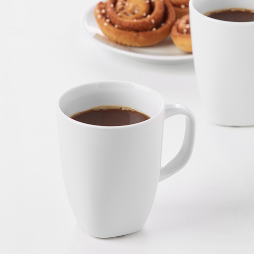 Dos tazas de cerámica blanca llenas de café sobre una mesa blanca, acompañadas de un plato de bollos de canela.