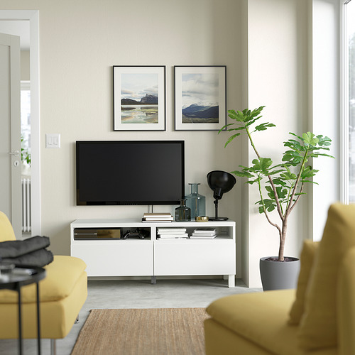 Sala de estar moderna con mueble de TV blanco BESTÅ, TV de pantalla plana, sillas amarillas, planta de interior y obras de arte enmarcadas. Diseño limpio y minimalista.