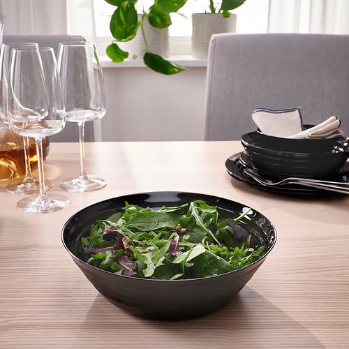 Cuenco negro con ensalada sobre mesa de madera, copas de vino, platos y servilletas.